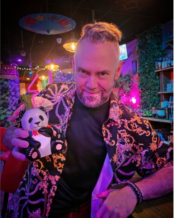 Dave behind the bar in an embroidered jacket, holding a panda tiki mug