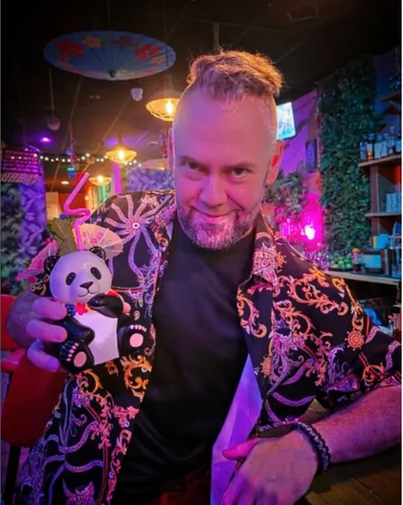 A bartender at The Tipsy Skipper in an embroidered jacket holding a panda tiki mug