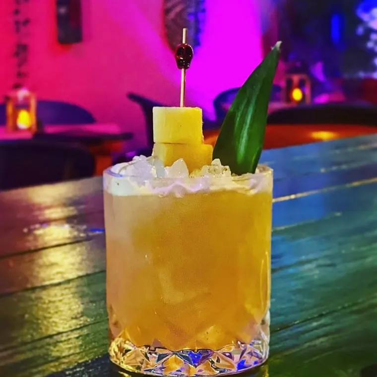A tall golden cocktail over crushed ice with a pineapple frond, lit by magenta neon