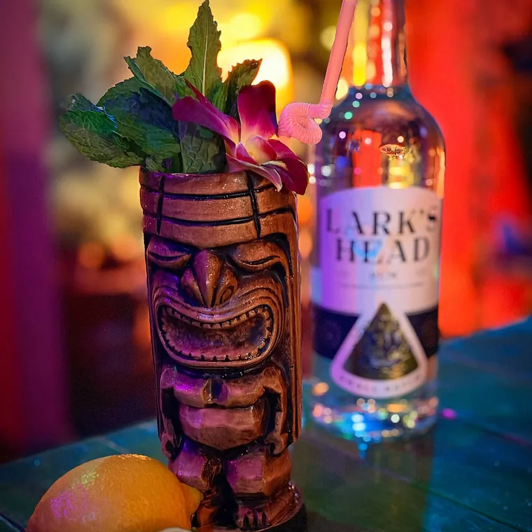 A carved tiki mug cocktail beside a bottle of Lark's Head rum and fresh grapefruit