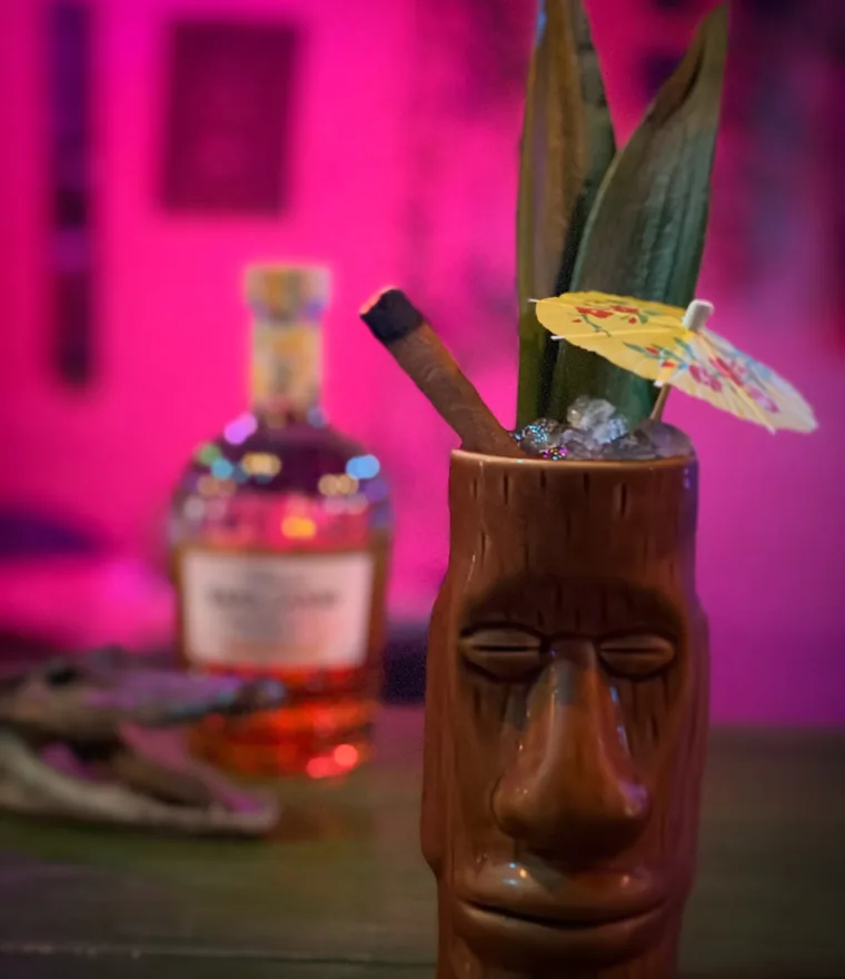 A cocktail in a carved wooden tiki mug with a paper parasol and pineapple frond, under magenta neon at The Tipsy Skipper in Ocala